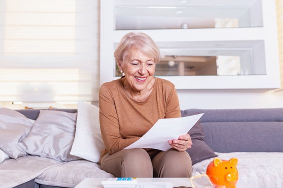 Femme indépendante senior souriante lisant des documents sur la retraite, avec une tirelire en forme de cochon à côté.